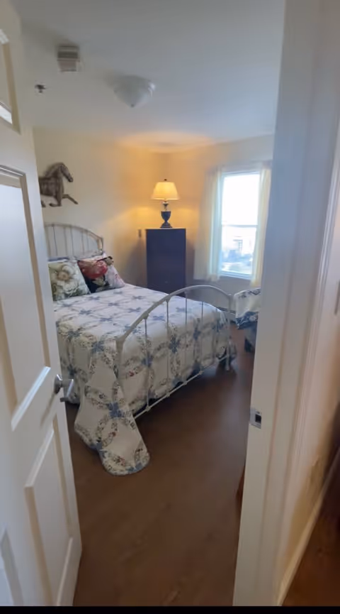 A small bedroom with a metal bed frame and a floral patterned quilt. There are several pillows on the bed, a wooden dresser with a lamp on top, and a window with white curtains letting in natural light. The floor is wooden, and the walls are painted a light color.