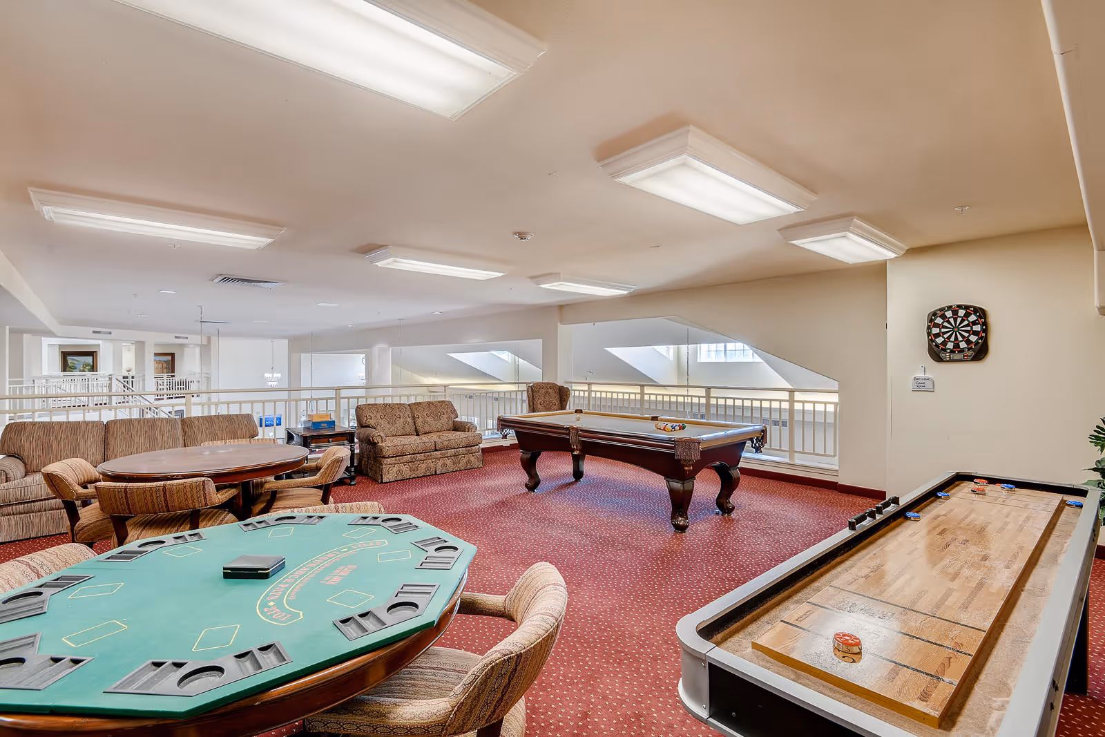 A recreational room with a poker table, a shuffleboard table, a pool table, and comfortable seating including sofas and chairs. The room has red carpet flooring, white walls, and ceiling lights. A dartboard is mounted on one wall.