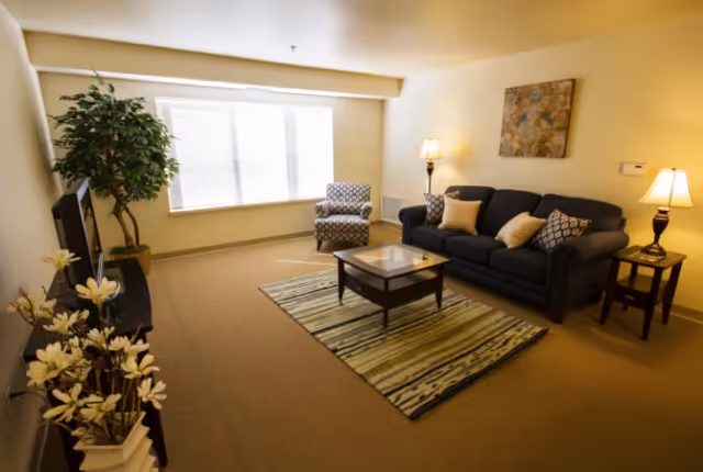 A cozy living room with a dark blue sofa adorned with several pillows, a patterned armchair, a glass-top coffee table on a striped rug, two side tables each with a lamp, a large window with blinds, a potted plant, and a piece of artwork hanging on the wall.