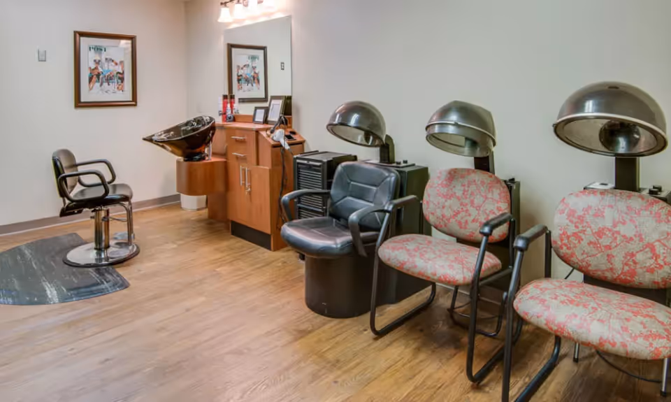 Interior of a hair salon area with wooden flooring, featuring a black salon chair in front of a wash basin, a wooden cabinet with a mirror above it, and two floral-patterned chairs with hair dryers attached behind them.