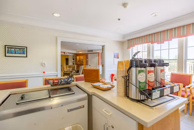 A coffee and beverage station with dispensers and paper cups in a bright senior living common area with chairs and a seating area beyond.