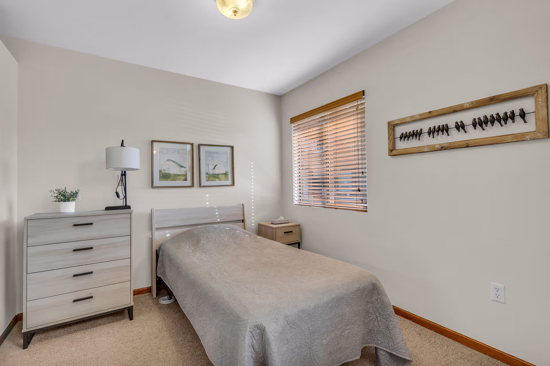 A simple single bedroom with a twin bed, dresser, nightstand, lamp, window blinds, and framed wall art.