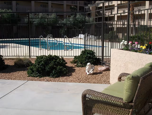 Wicker patio chair and planter in a courtyard beside a fenced swimming pool with apartment balconies in the background.