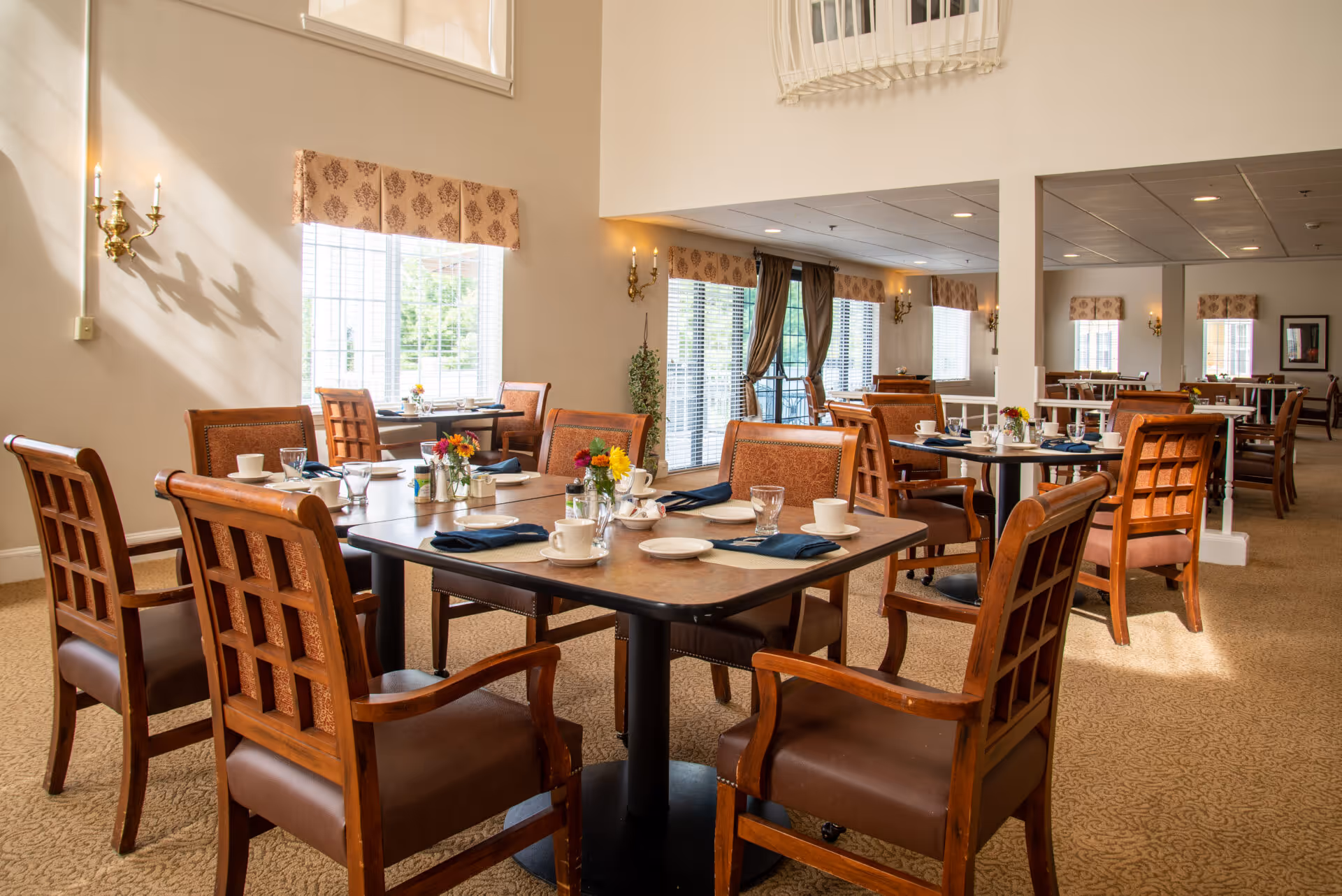 Bright, spacious dining room with wooden tables and chairs set for meals and windows letting in natural light.