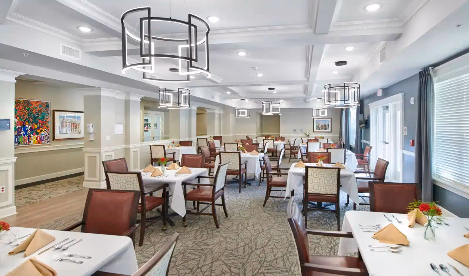 A bright and spacious dining room with multiple tables covered in white tablecloths, each set with beige napkins, silverware, and small floral centerpieces. The room features modern circular pendant lights hanging from a coffered ceiling, patterned carpet flooring, and large windows with blinds and curtains allowing natural light to enter. Artwork is displayed on the walls, and the overall decor is elegant and welcoming.