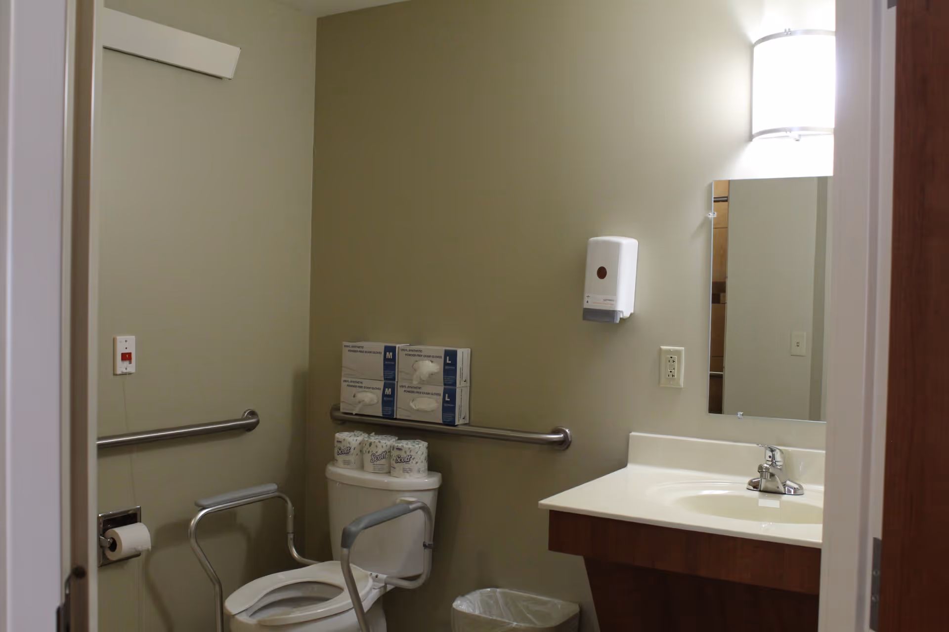 Accessible bathroom with a toilet fitted with safety rails, grab bars, a sink and mirror, wall-mounted dispenser, and stocked supplies.