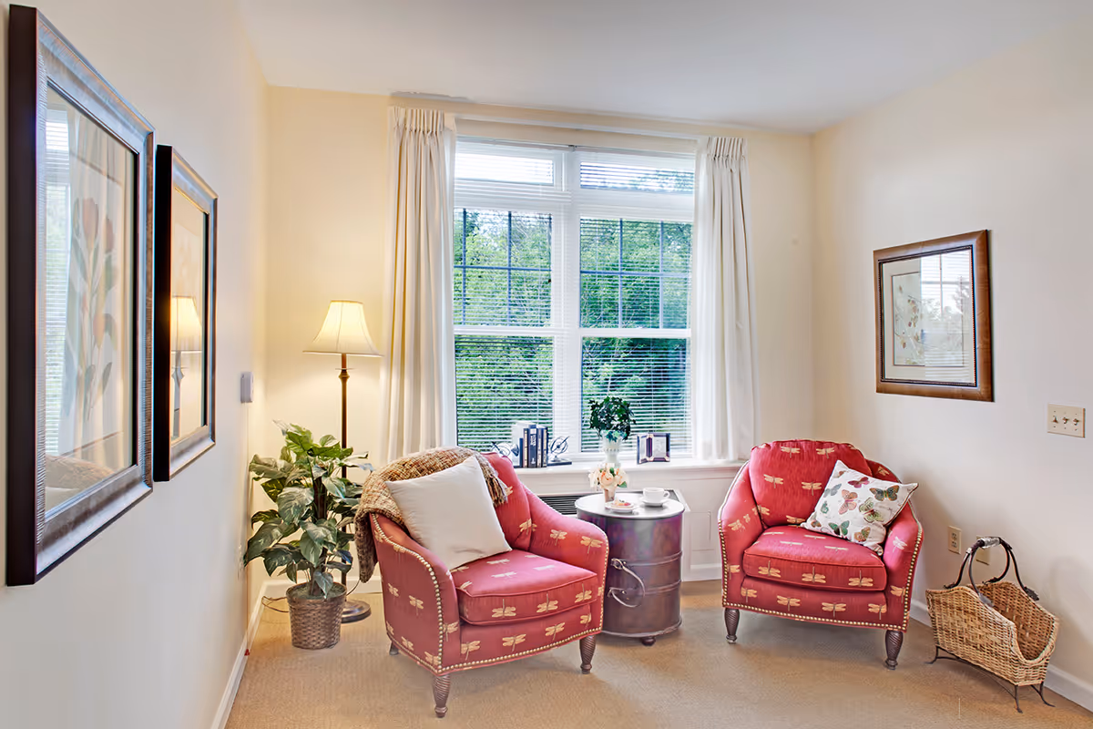 Sunlit sitting area with two red upholstered armchairs, a small round table, floor lamp, framed art, and a large window overlooking greenery.