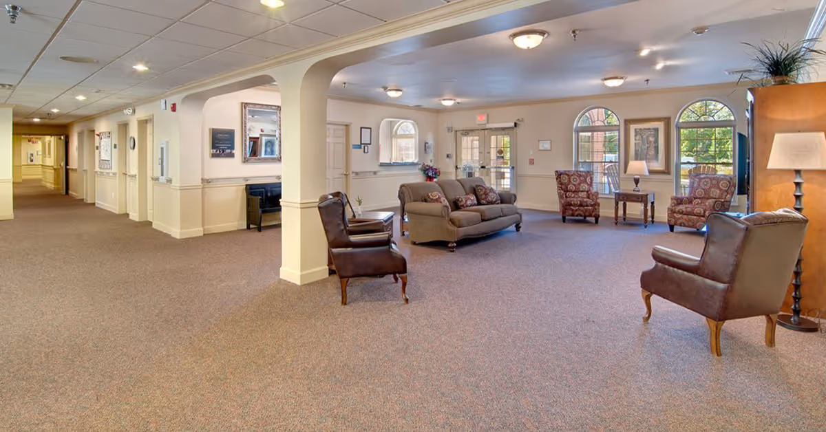 Spacious furnished common lounge in a senior living facility with sofas, armchairs, lamps and arched windows.