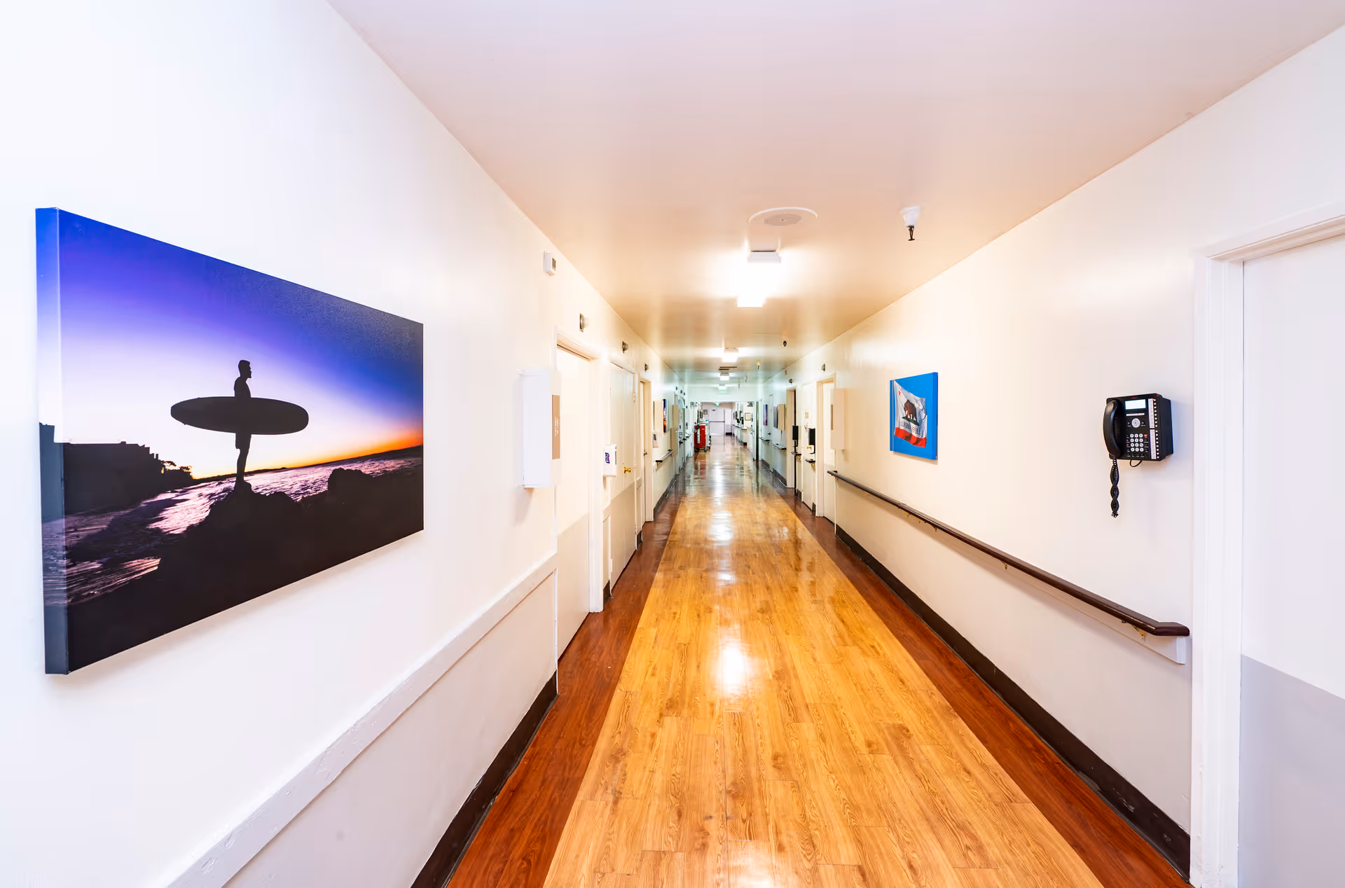 A long, well-lit hallway with wooden flooring and white walls. The hallway has handrails on both sides and several doors along the walls. There are two framed pictures hanging on the walls, one showing a silhouette of a person with a surfboard at sunset and another smaller colorful picture. A black wall-mounted telephone is visible on the right side.