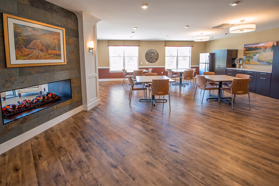 A spacious dining room with several tables and chairs arranged on a wooden floor. The room features large windows allowing natural light, a modern electric fireplace with a framed painting above it, and cabinetry with a refrigerator and coffee machine. The walls are painted in warm tones with decorative wall art.