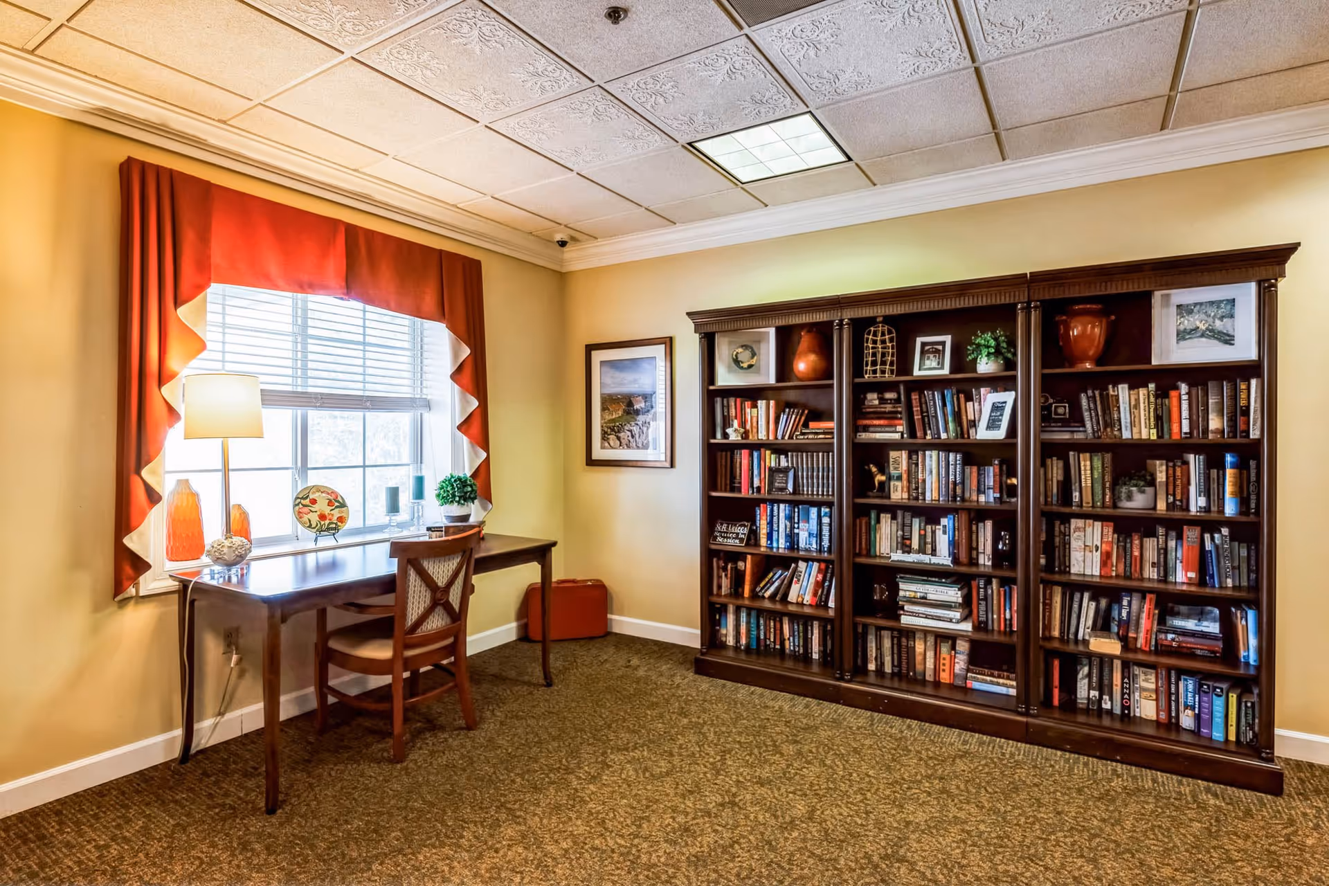 A cozy interior room with a large wooden bookshelf filled with books and decorative items on the right side. On the left side, there is a wooden desk with a chair in front of a window with red and white curtains. The desk has a lamp, a decorative plate, and a small plant. The walls are painted light yellow, and the floor is carpeted in a patterned brown color.