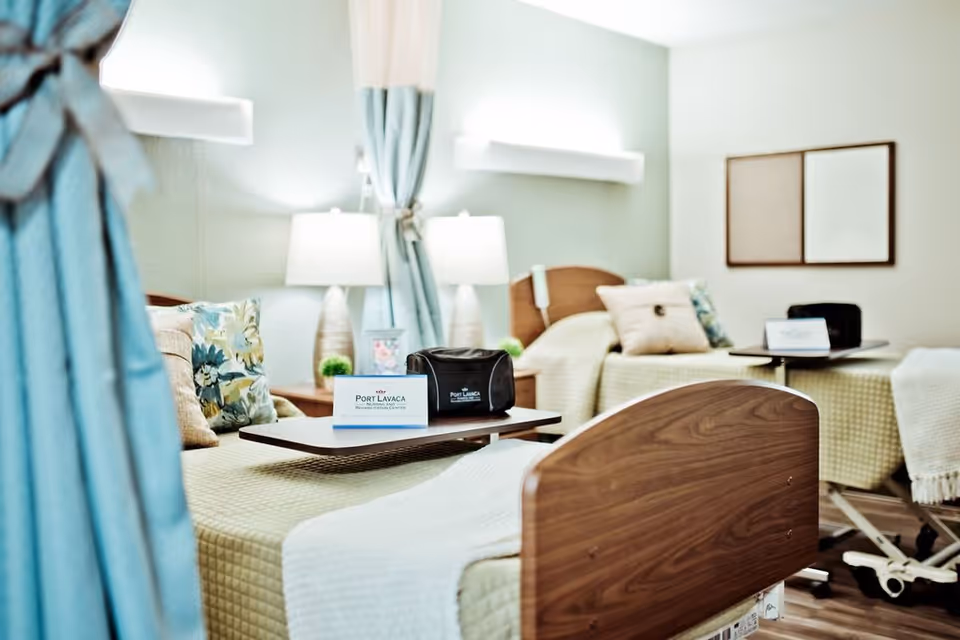 A bright and clean nursing facility room with two hospital-style beds, each with a beige quilted blanket and decorative pillows. Each bed has a small overbed table with a Port Lavaca Nursing and Rehabilitation Center card and a black bag on top. There are two bedside lamps and light blue curtains partially drawn around the beds. The walls are light-colored with a simple framed artwork.