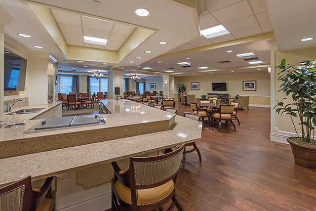 Spacious senior living facility common area with a large countertop bar, multiple tables and chairs, wood flooring, and soft yellow walls. The room is well-lit with recessed lighting and natural light from windows. There are plants and framed pictures on the walls, creating a welcoming atmosphere.