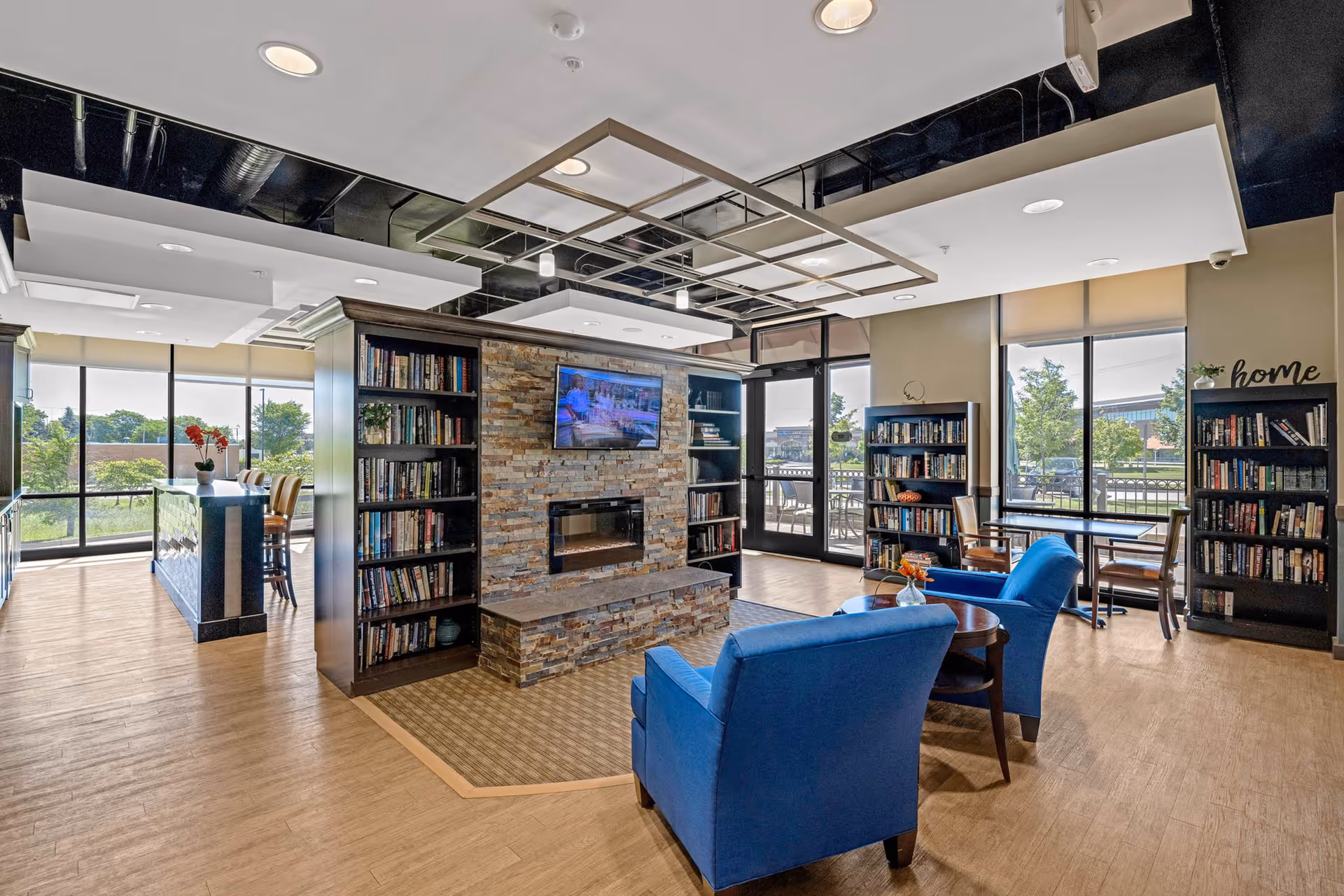 A cozy common area in a senior living facility with large windows letting in natural light. The room features a stone fireplace with a mounted TV above it, surrounded by bookshelves filled with books. There are two blue armchairs and a small round table in front of the fireplace. Additional seating and tables are visible near the windows, and a kitchen or bar area is seen to the left with high chairs. The space has a modern ceiling design with recessed lighting.