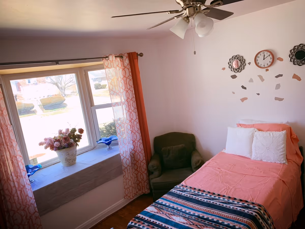 Bright bedroom with a single bed covered in pink linens and a patterned blanket, an armchair, and a window seat with curtains and a vase of flowers.