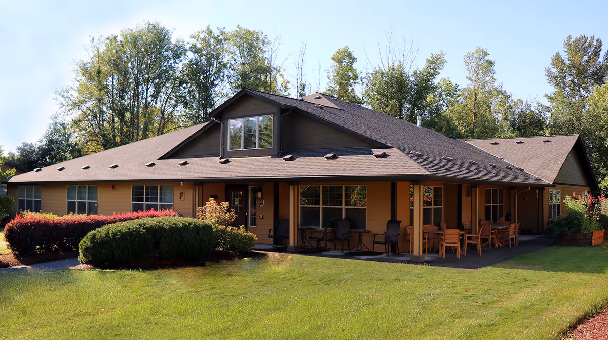 Exterior view of a single-story senior living facility building with a sloped roof, surrounded by green grass, bushes, and trees under a clear blue sky. The building has a covered porch area with outdoor seating including tables and chairs.