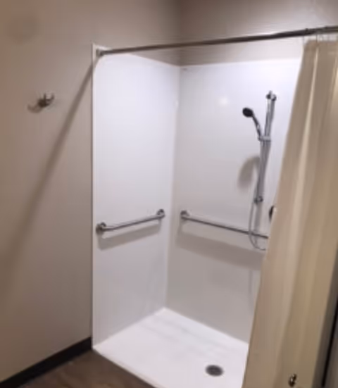 A clean, white walk-in shower with two horizontal grab bars on the walls and a handheld showerhead mounted on a vertical bar. A beige shower curtain is partially drawn to the right side. The floor outside the shower is wood-toned, and there is a single wall hook on the left wall.
