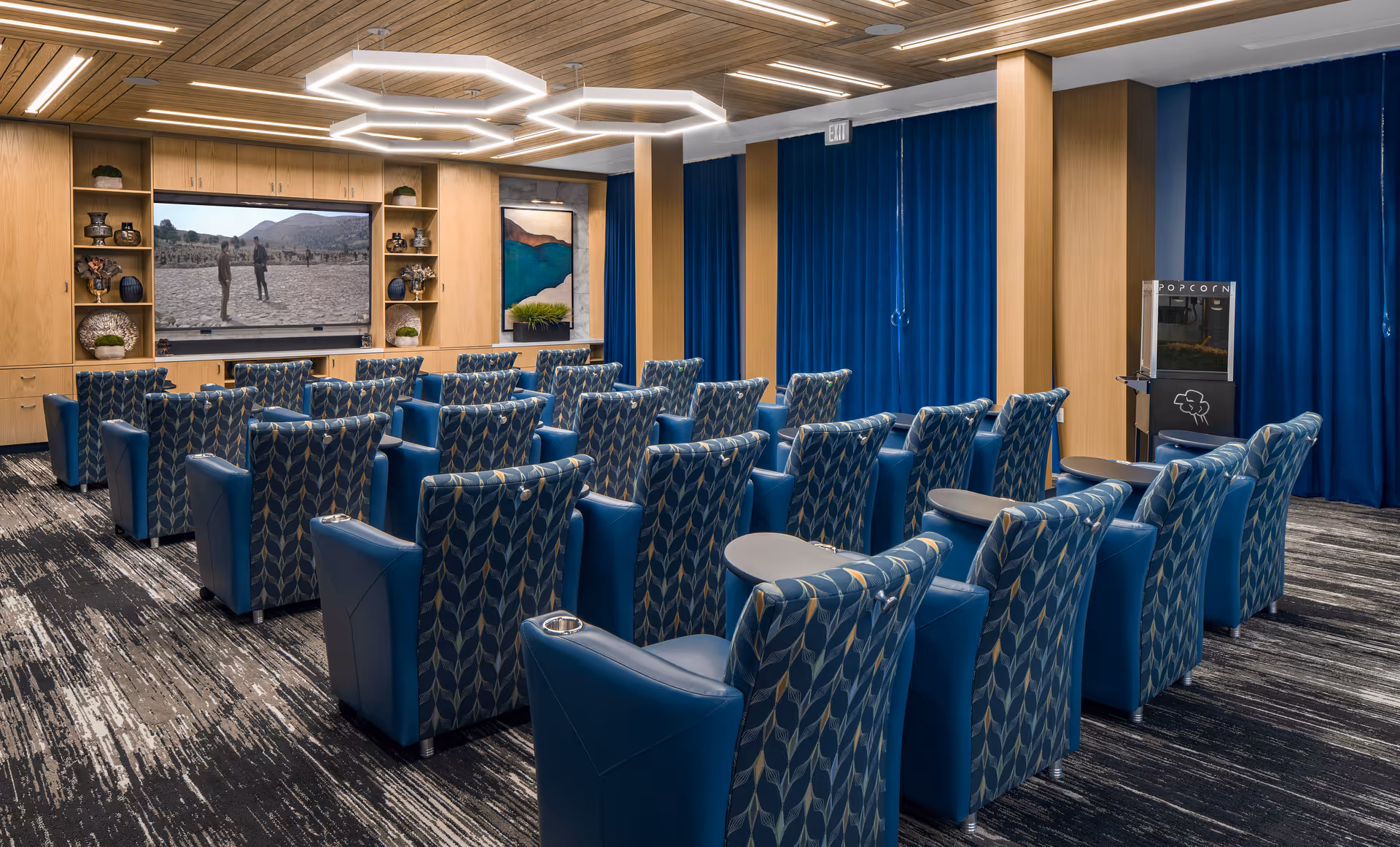 A modern media room with multiple rows of blue patterned armchairs facing a large flat-screen TV mounted on a wooden wall unit. The room features a popcorn machine on the right side, blue curtains along the walls, and geometric ceiling lights. The carpet has a textured black and white pattern.