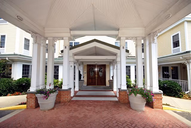 Covered entrance with white columns, potted flowers, and double wooden doors at a yellow multi-story building.