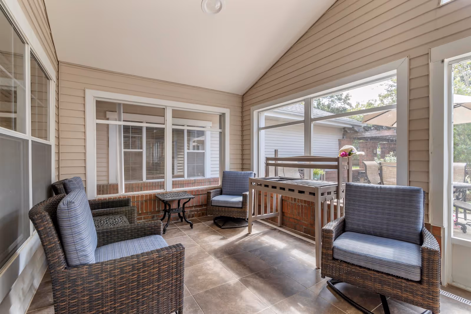 A sunroom with large windows on two sides, featuring four wicker chairs with blue cushions arranged around a small black metal table. There is a wooden bench with a flower pot on top against the wall. The room has beige walls with white trim and a tiled floor.