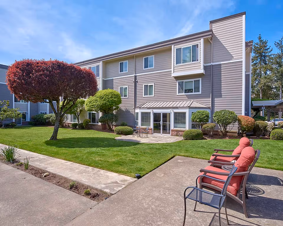 Outdoor view of a senior living facility building with neatly trimmed bushes and trees, a green lawn, a concrete patio with three cushioned chairs, and a small table with two chairs near the building entrance under a clear blue sky.