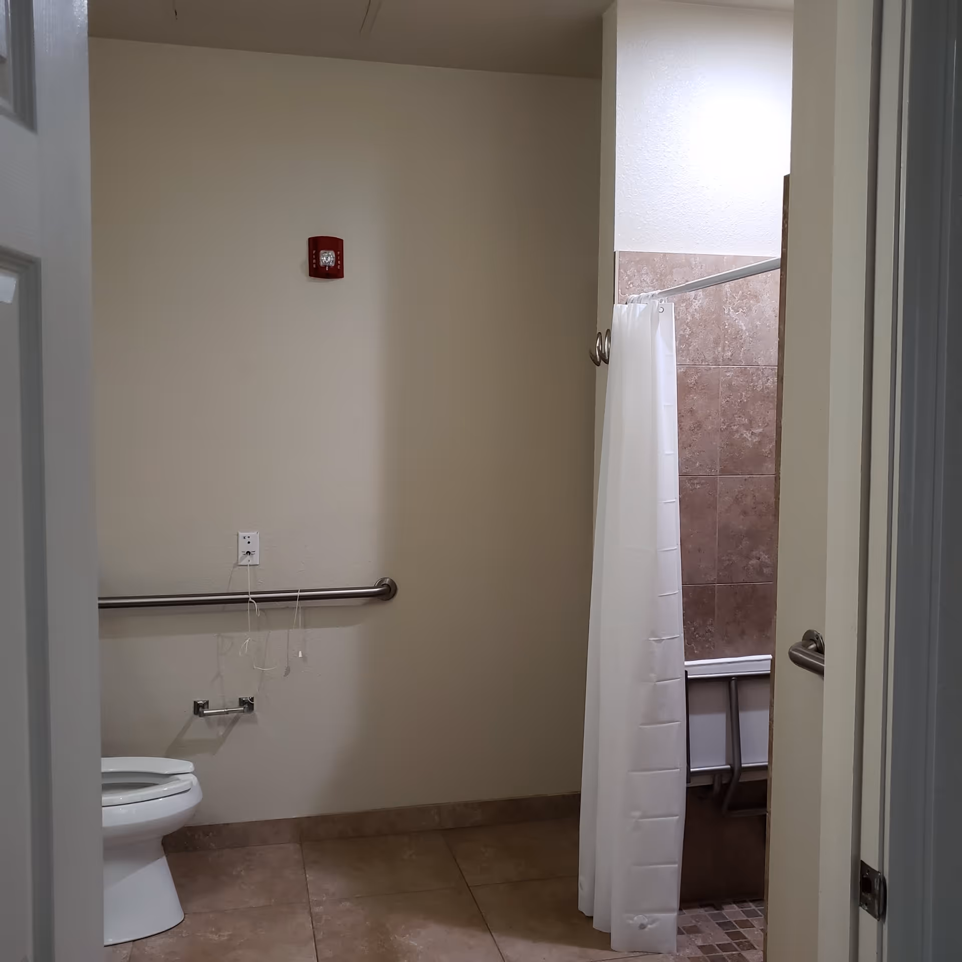 A bathroom with a white toilet on the left side, beige tiled floor, and a shower area with a white curtain and brown tiled walls on the right. There are metal grab bars on the walls for support and a red fire alarm on the wall above the toilet.