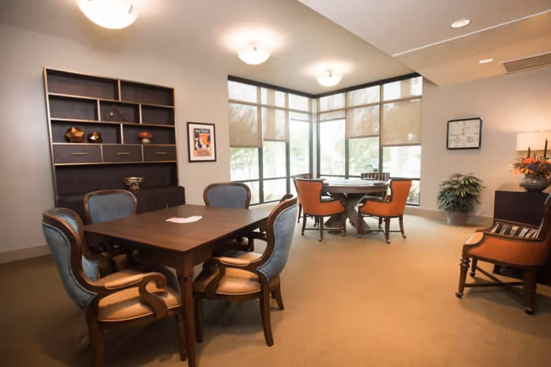 A well-lit room with two wooden tables surrounded by chairs. One table has six blue cushioned chairs, and the other round table has four orange cushioned chairs. The room has large windows with beige blinds, a dark wooden shelving unit with decorative items, a framed picture on the wall, a potted plant, and a side table with a lamp and floral arrangement.