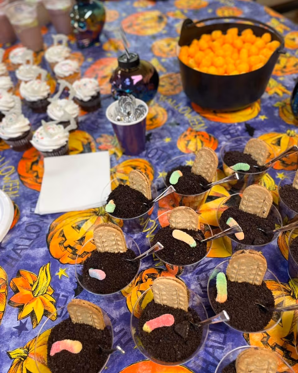 A Halloween-themed table setting with dessert cups filled with crushed cookies resembling dirt, topped with gummy worms and a cookie resembling a gravestone. There are also cupcakes with white frosting and sprinkles, a black cauldron filled with cheese balls, and a purple cup holding spoons. The tablecloth features pumpkins and Halloween decorations.
