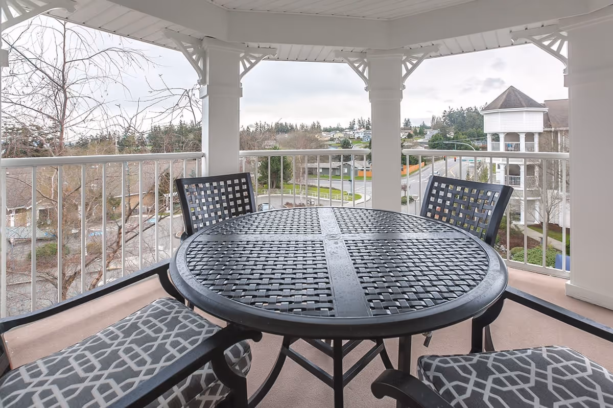 Covered outdoor balcony with a round black metal table and four chairs, two with patterned cushions. The balcony overlooks a street, trees, and nearby buildings under a cloudy sky.
