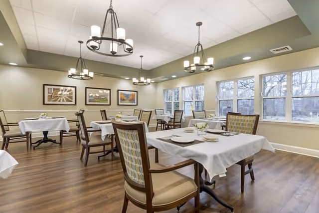 Bright dining room with multiple tables set with white tablecloths and chairs under chandeliers and large windows.
