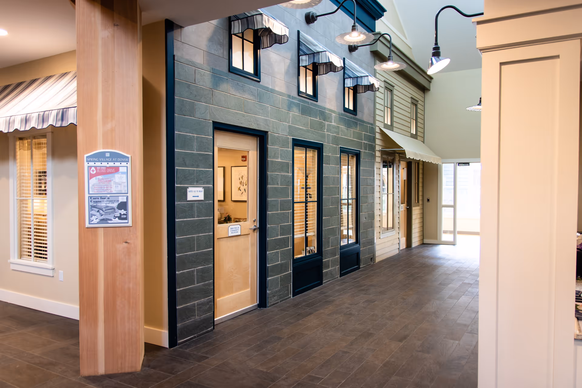 Indoor hallway designed to resemble an outdoor street with building facades, windows with awnings, and doors. The floor is tiled, and there are wall-mounted lights above the windows. A wooden pillar on the left has a bulletin board with notices, and there is a glass door at the end of the hallway letting in natural light.