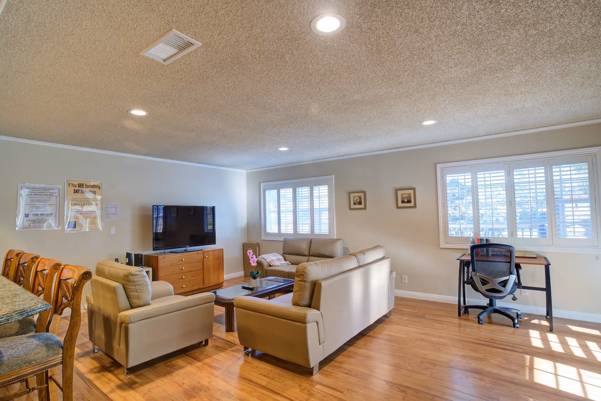A bright living room with beige leather sofas arranged around a wooden coffee table, a flat-screen TV on a wooden cabinet, a desk with a chair near windows with white shutters, and wooden bar stools at a counter on the left side.