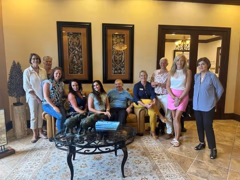 A group of nine adults, five women and one man seated on a tan couch and three women standing behind and beside the couch, in a warmly lit living room with beige walls, decorative framed artwork, a glass coffee table with a sculpture of three elephants, and a patterned rug on the tiled floor.
