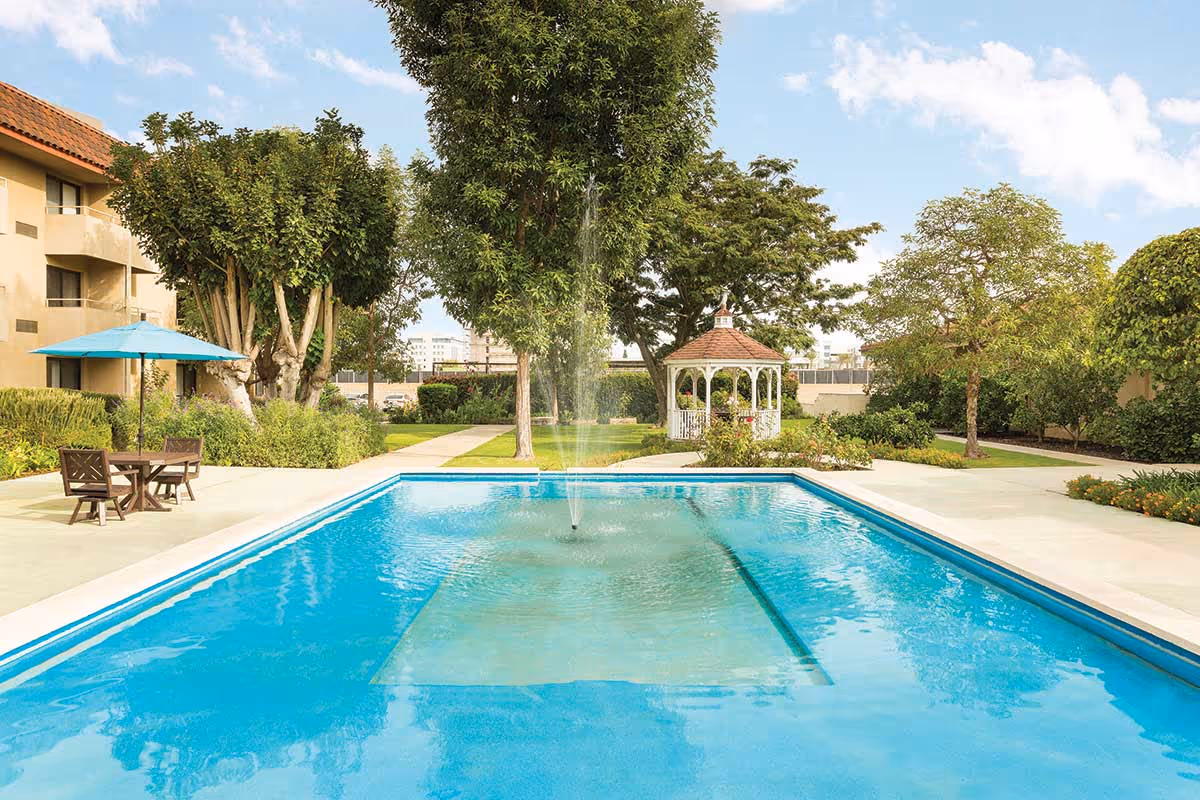 Outdoor swimming pool with a central water fountain, surrounded by a concrete deck with a table, chairs, and an umbrella on the left side. In the background, there are trees, bushes, a white gazebo, and a multi-story building with balconies.