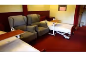 A nursing center room with two hospital beds, two gray recliner chairs, and overbed tables. The room has red carpet flooring and yellow walls with a window covered by a red curtain.