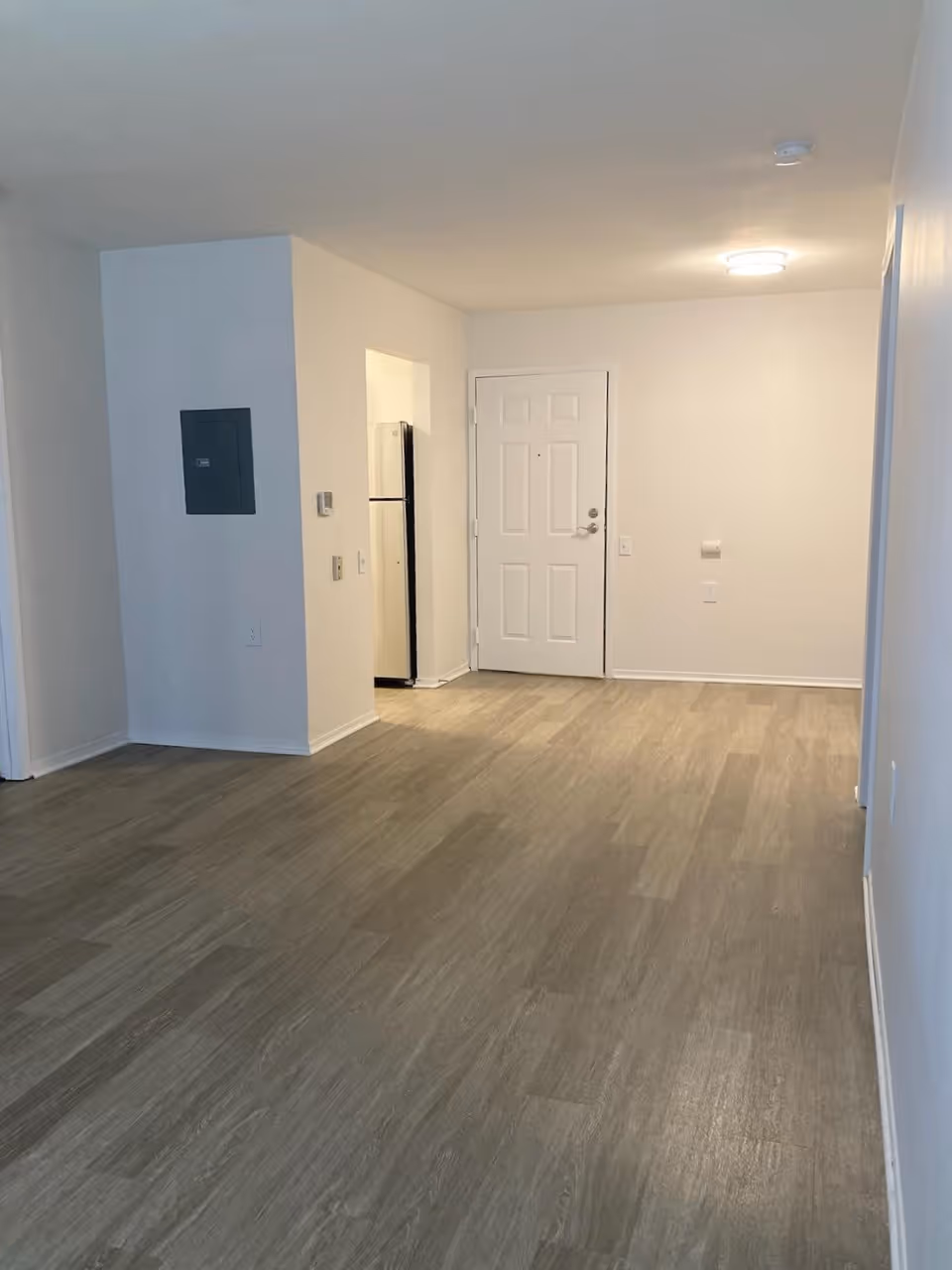 Empty interior space with light wood flooring, white walls, a white front door, and a small kitchen area visible with a refrigerator.