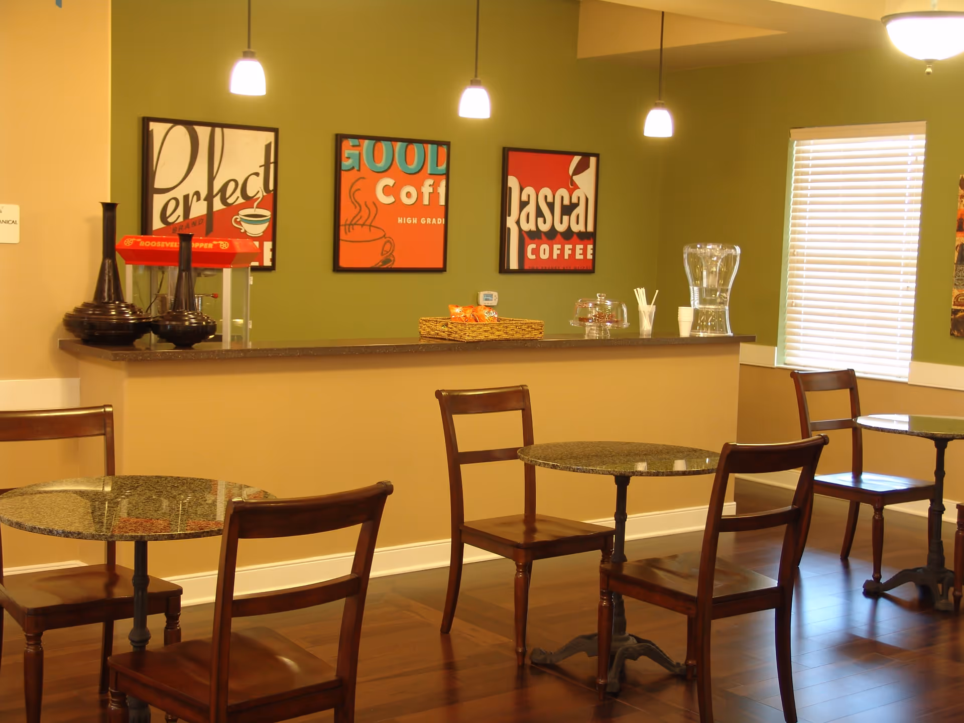 A cozy dining area with small round tables and wooden chairs. A counter with a popcorn machine, decorative vases, a basket of snacks, a glass water pitcher, and cups is against a green wall. Three framed coffee-themed posters hang on the wall above the counter. Pendant lights hang from the ceiling, and a window with blinds is on the right side.