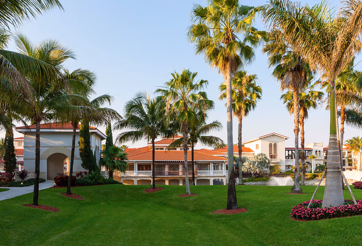 Front view of a Mediterranean-style senior living complex with palm trees, manicured lawn, a fountain, and red-tiled roof buildings.