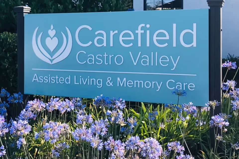 A blue sign with white text reading 'Carefield Castro Valley Assisted Living & Memory Care' surrounded by purple flowers and greenery.