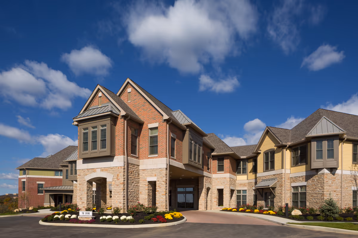 Brick-and-stone senior living building with arched entrance and landscaped circular drive under blue sky