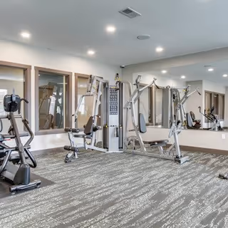 A fitness room with various exercise machines including a stationary bike, weight machines, and other gym equipment. The room has large windows, mirrors on the walls, and recessed ceiling lights. The floor is carpeted with a patterned design.