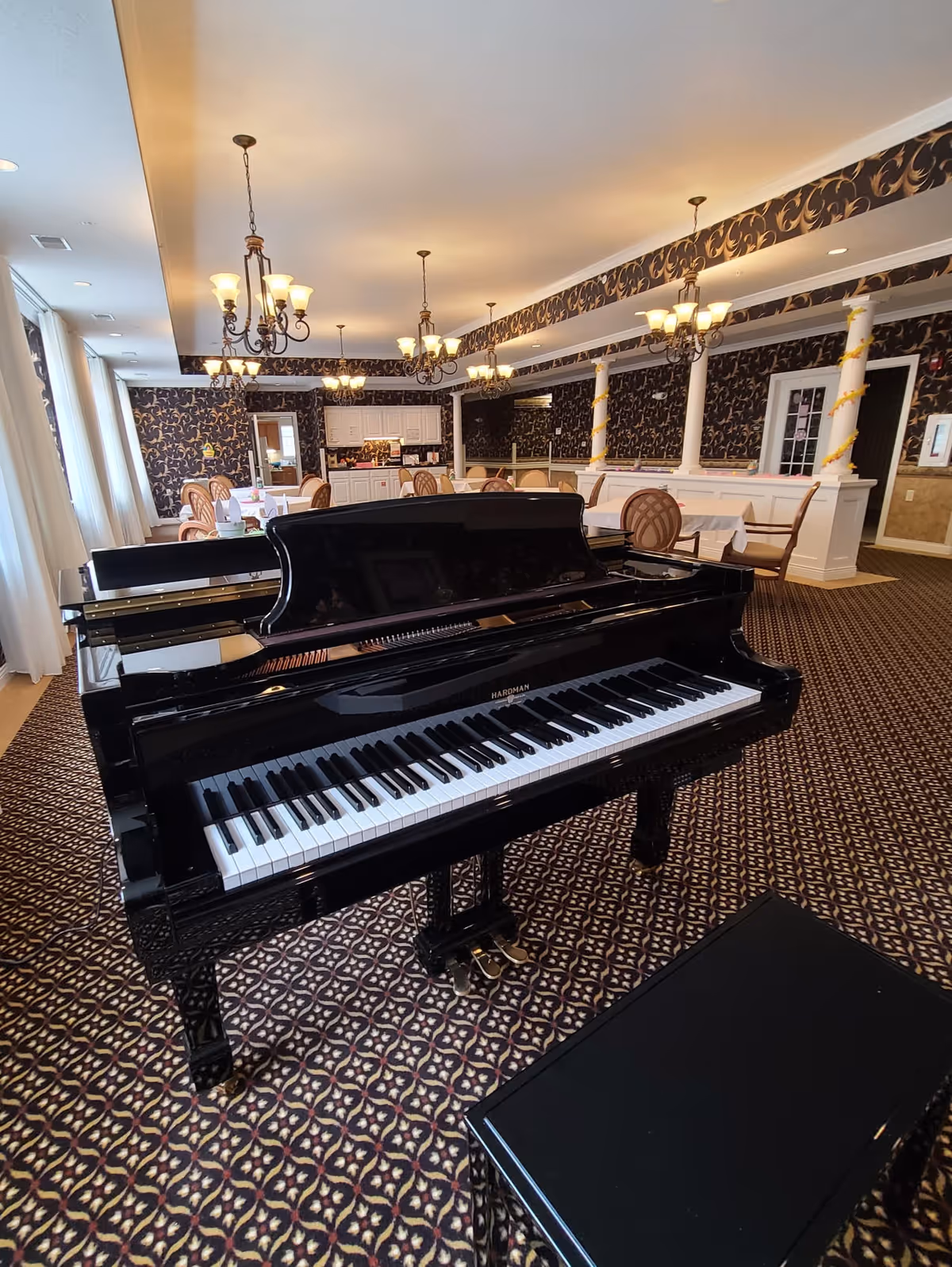 Interior view of a dining room in Kingston Court Assisted Living featuring a black grand piano in the foreground. The room has patterned carpet, ornate chandeliers, and tables with chairs arranged for dining. The walls are decorated with dark floral wallpaper and white columns wrapped with gold ribbon accents.