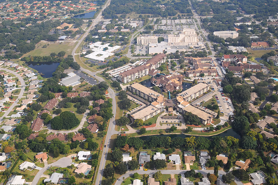 Aerial view of a residential area featuring a large senior living facility with multiple connected buildings arranged in a star-like pattern, surrounded by trees, roads, and smaller houses. The facility is identified as Freedom Village of Bradenton.