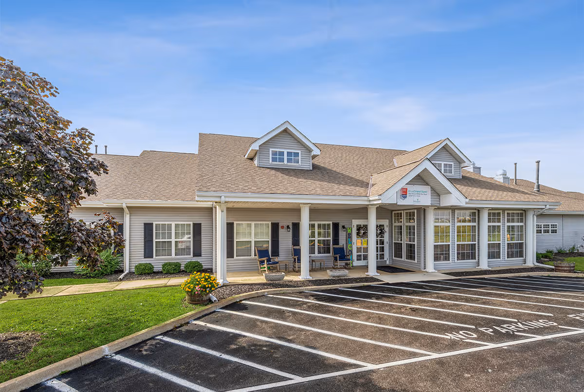 Exterior view of Digby Place senior living facility showing a single-story building with a covered entrance, large windows, and a parking lot with marked spaces. There are some shrubs and a tree on the left side, and a clear blue sky above.