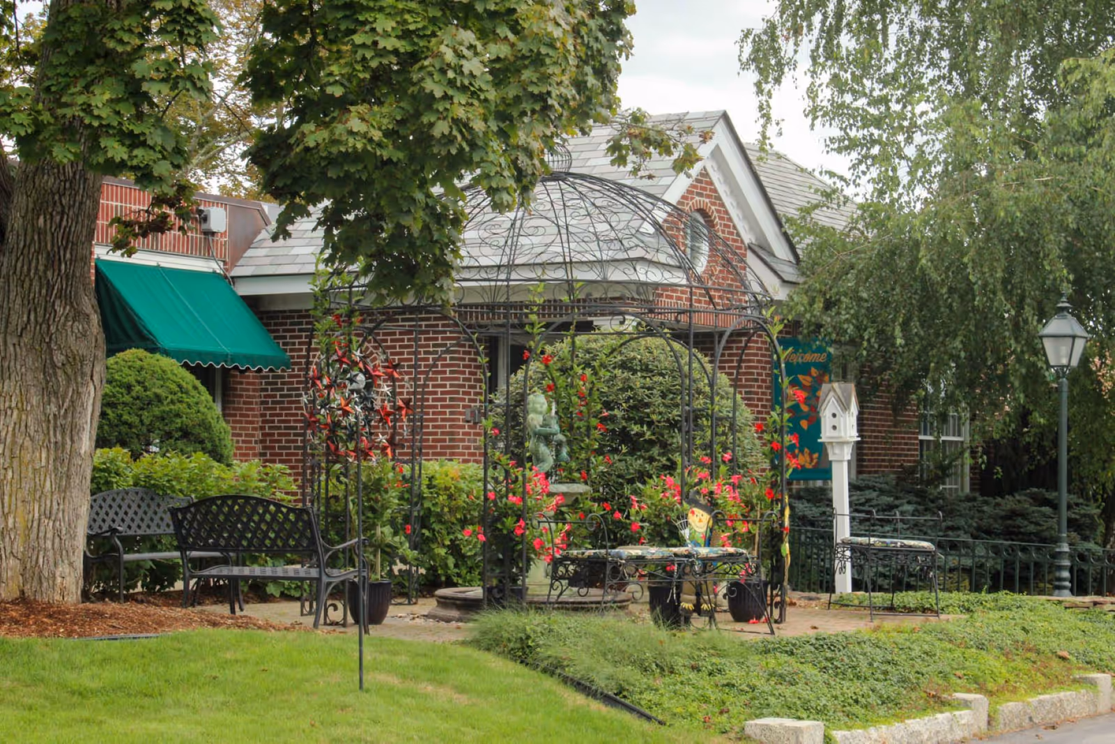Front garden of a brick building featuring a wrought-iron arbor with flowers, benches, a birdhouse post and a green awning.