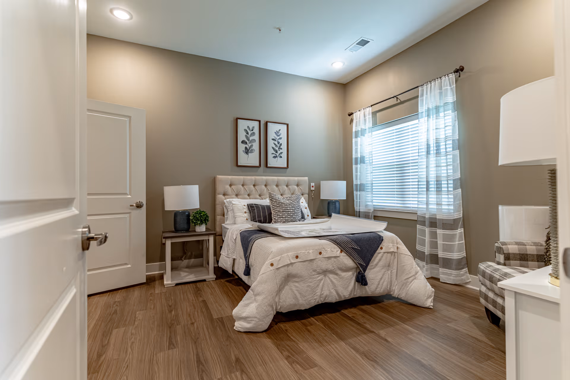 A cozy bedroom with a neatly made bed featuring white and gray bedding, two framed botanical prints on the wall above the tufted headboard, two bedside tables each with a lamp and a small plant on one, a window with sheer striped curtains allowing natural light, and a checkered armchair in the corner.