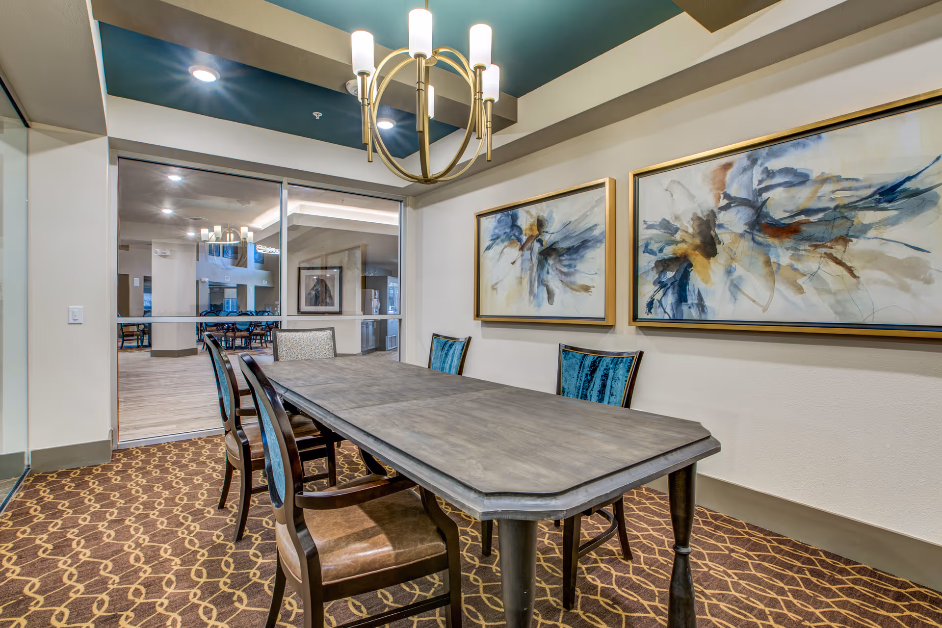 A small dining area with a rectangular wooden table and six chairs, some with blue upholstery. The room features a patterned carpet, two abstract paintings on the wall, and a modern chandelier hanging from a recessed ceiling. A glass partition separates this room from a larger dining area visible in the background.