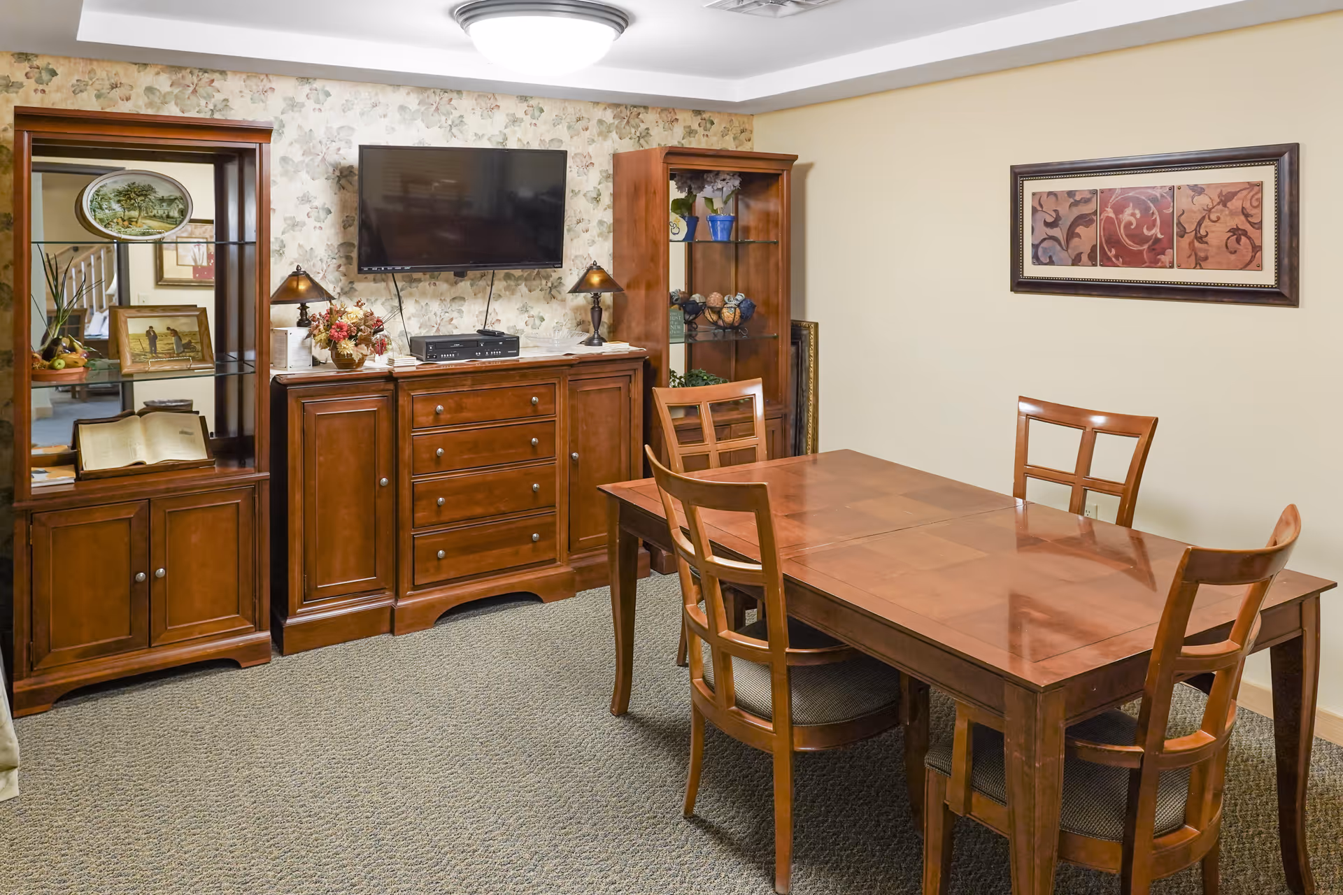 A dining room with a wooden dining table and four chairs. Against the wall is a wooden cabinet with drawers and two glass display cabinets on either side. A flat-screen TV is mounted above the cabinet. The walls are decorated with floral wallpaper and a framed artwork. The floor is carpeted.