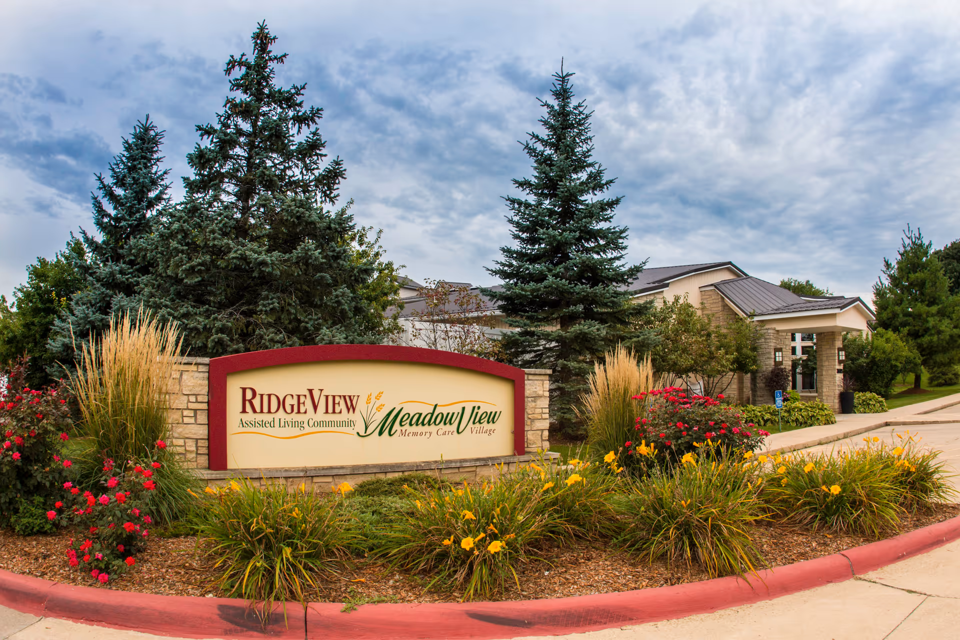 Entrance sign for RidgeView Assisted Living Community and MeadowView Memory Care Village surrounded by landscaped flowers and trees, with the facility building visible in the background under a cloudy sky.