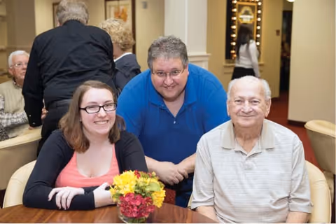 Three people sitting and standing around a table with a small flower arrangement in a well-lit indoor setting. Two elderly men and a younger woman are smiling at the camera, with other people and furniture visible in the background.